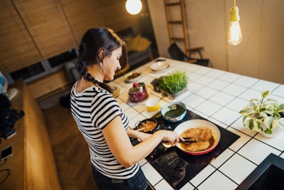 Young woman cooking a healthy meal in home kitchen.Making dinner on kitchen island standing by induction hob.Preparing fresh meal,enjoying spice aromas.Eating in.Passion for cooking.Healthy lifestyle and dieting concept.