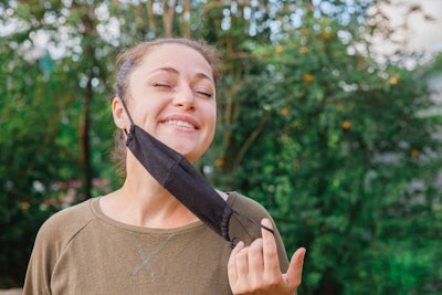 Happy positive girl takes off protective medical mask from face outdoors. Young woman removing mask smiling after vaccination. Coronavirus pandemic Covid 19 concept. Spring flowers pollen allergy