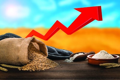 Front view of a sack of wheat laying down on a wooden table beside a bowl and a serving scoop fool of flour. Behind the table is a defocused wheat field and a moving up red arrow. This image represents the increase in the price of wheat caused by the conflict between Ukraine and Rusia.