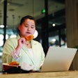 A plump young asian woman, dressed in a green suit, eats hamburgers while working on a laptop in a fast food restaurant.